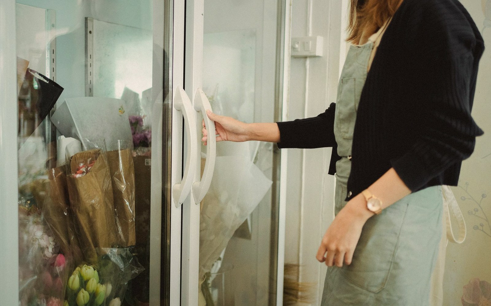 A woman opens a glass refrigerator door filled with wrapped flower bouquets